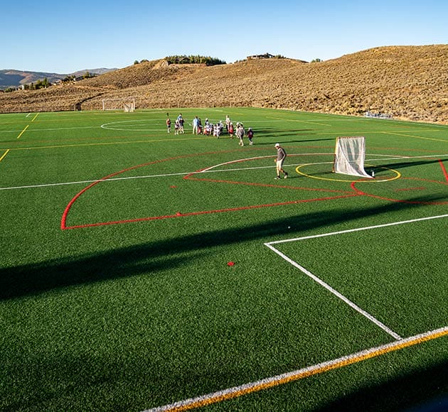 Artificial turf sports field with players and a lacrosse goal post.